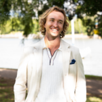 Man with wavy blond hair in a cream blazer smiles outdoors by a lake, with trees in the background.