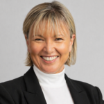 Smiling professional woman in a black blazer with arms crossed, standing against a light gray backdrop.