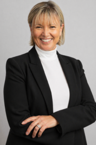 Professional woman with short blonde hair in a black blazer and white turtleneck, smiling with arms folded against a neutral gray background